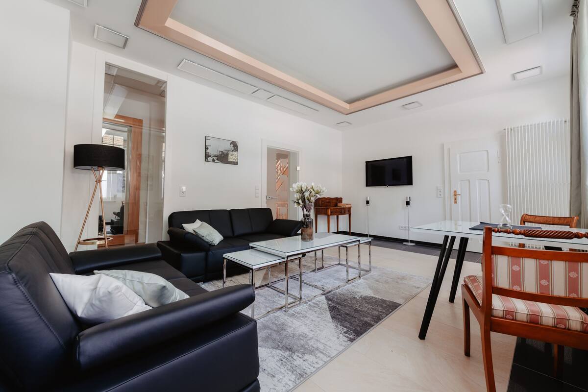 A modern living room with a black leather sofa, a glass coffee table, and a wooden dining table with chairs.