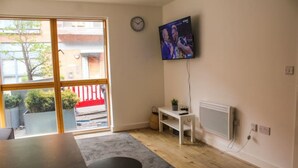Et værelse med et stort vindue, et fladskærms-tv monteret på væggen, et lille bord og en radiator.