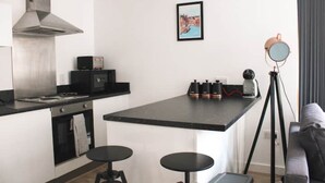 Et moderne køkken med en central ø, indbygget ovn og et emhænde i rustfrit stål. Der er to sorte barstole og et lille bord ved øen.
