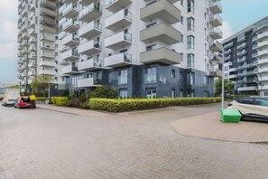 A modern apartment building with multiple balconies, a paved driveway, and a green trash bin.
