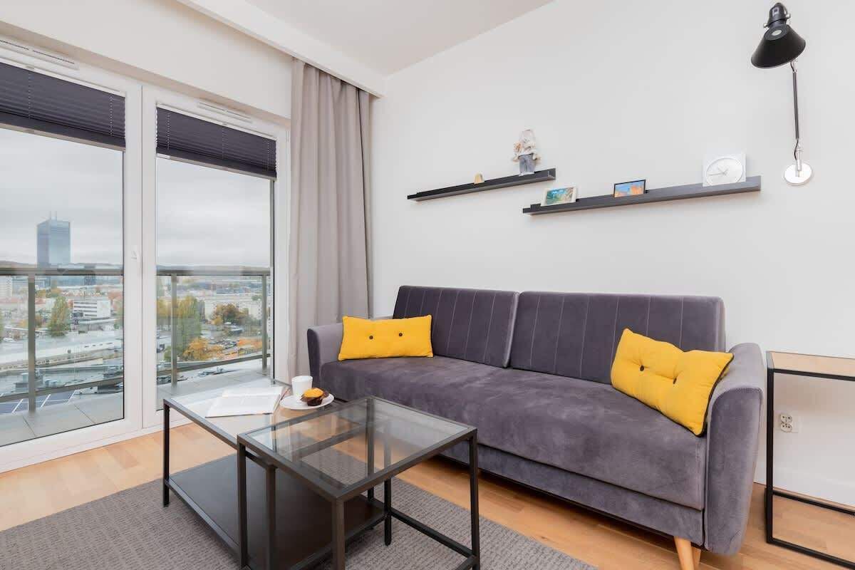 Uma sala de estar moderna com um sofá cinzento, almofadas amarelas, uma mesa de centro de vidro e vista para a cidade por grandes janelas.