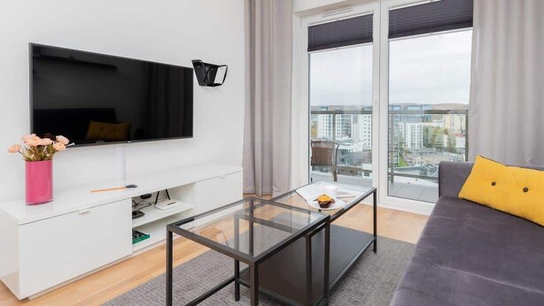 A modern living room with a flat-screen TV, a glass coffee table, a grey sofa, and a city view through large windows.