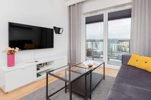 A modern living room with a flat-screen TV, a glass coffee table, a grey sofa, and a city view through large windows.