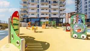 A playground with colorful play structures, including a green train and a yellow seesaw, in front of a modern apartment building.