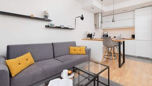 A modern living room with a grey sofa, yellow cushions, a glass coffee table, and a small bookshelf.