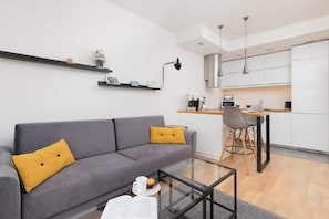 A modern living room with a grey sofa, yellow cushions, a glass coffee table, and a small bookshelf.