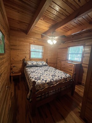 Un dormitorio de cabaña de madera con una cama, dos ventanas, un ventilador de techo y una mesita de noche pequeña.