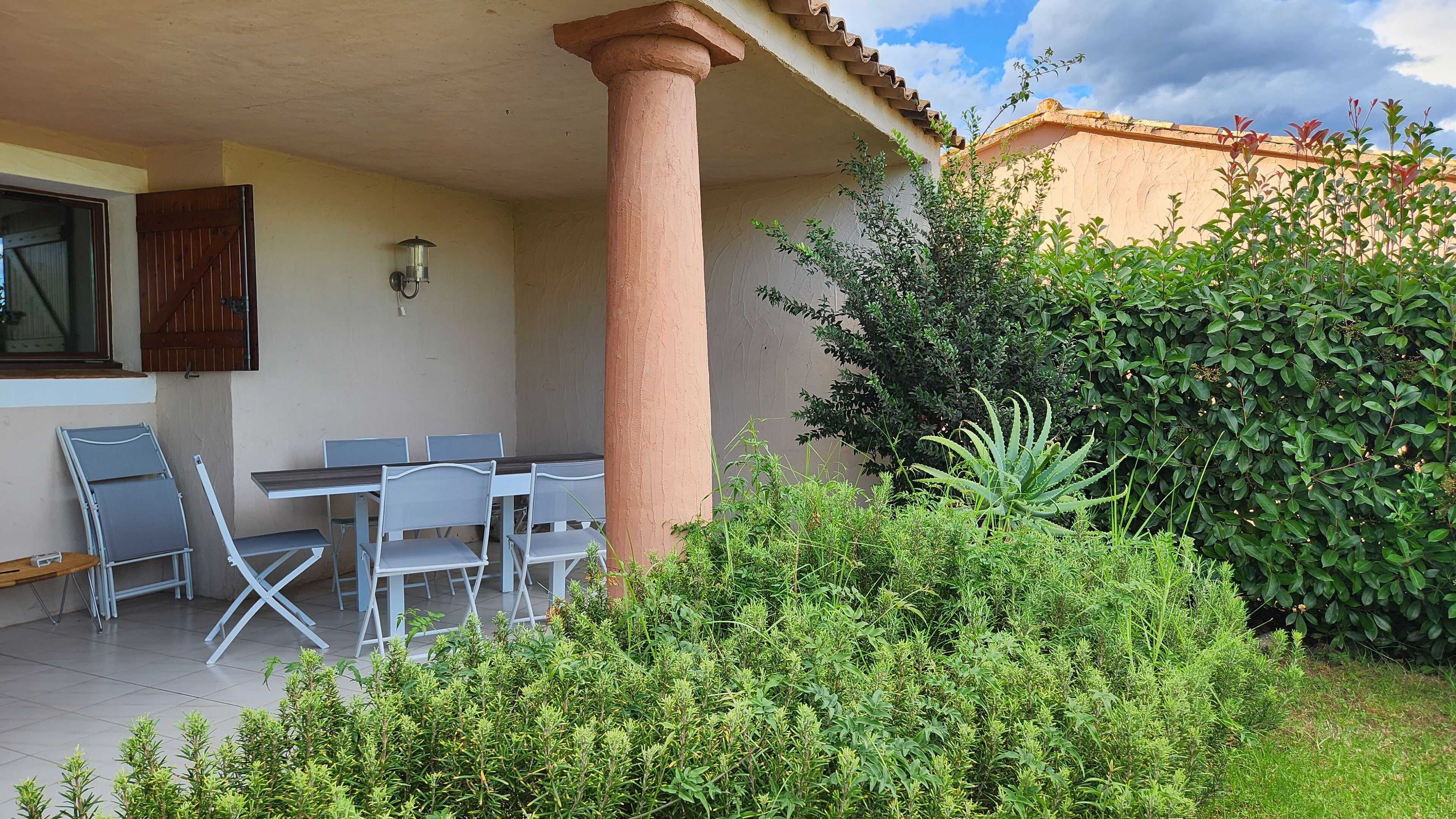 An outdoor patio with a table and chairs, surrounded by greenery and a building with a tiled roof.