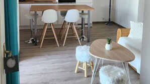 A modern kitchen with white cabinets, a wooden countertop, and a built-in oven. There is a dining area with a wooden table and white chairs. A round coffee table with a white fluffy stool is in the foreground. - A 250 m de la Plage de Favone, Appartement vue mer Avec Terrasse de 30m2 (Conca)