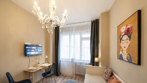A hotel room with a wooden floor, a desk with a chair, a bed with a patterned throw, a chandelier, a TV on the wall, and a large painting. - Old Town - Studio Petrin Tower (Prague)