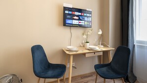 A modern room with a wooden desk, two blue chairs, a flat-screen TV mounted on the wall displaying various apps, a small potted plant, and a fan on the floor. - Old Town - Studio Petrin Tower (Prague)