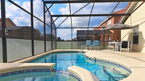 En swimmingpool med et buet design, omgivet af en overdækket patio med stole og parasoller.