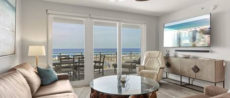 A modern living room with a sofa, a round coffee table, a TV mounted on the wall, and a balcony with a view of the beach.