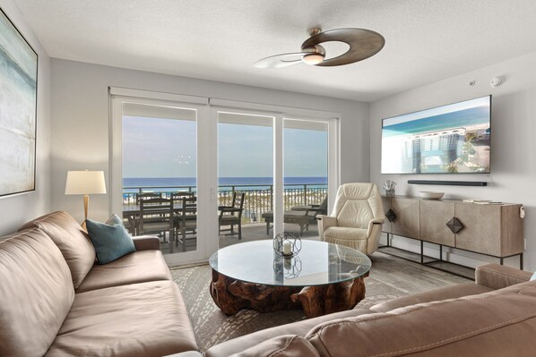 Een moderne woonkamer met een bank, een ronde salontafel, een aan de muur bevestigde televisie, en een balkon met uitzicht op het strand.