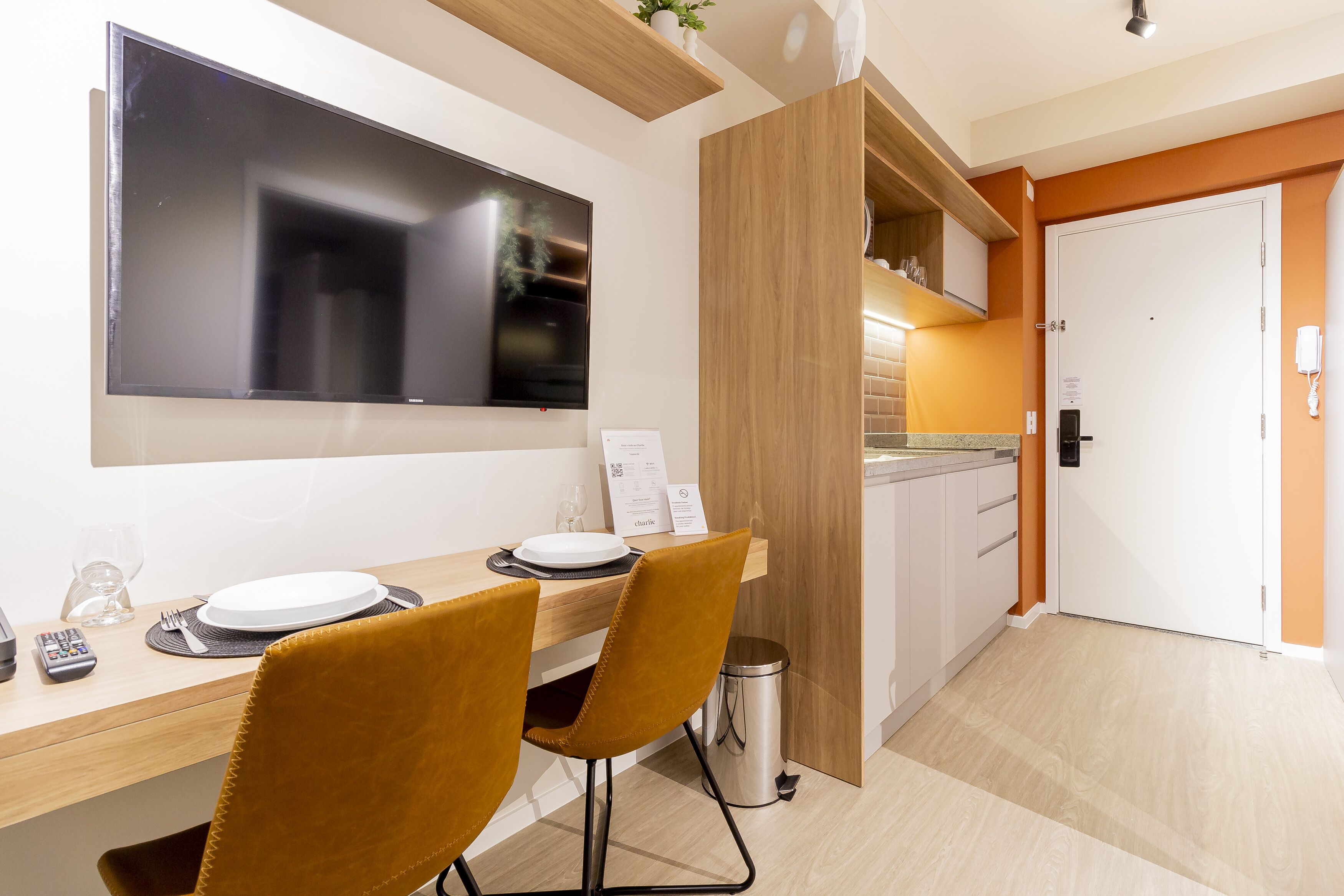 A modern dining area with a wooden table, two chairs, a TV mounted on the wall, and a kitchen area visible through an open door.