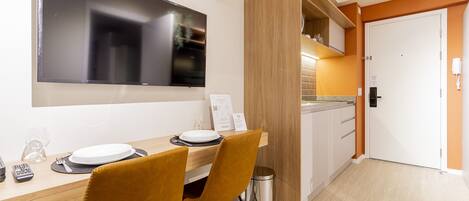 A modern dining area with a wooden table, two chairs, a TV mounted on the wall, and a kitchen area visible through an open door.