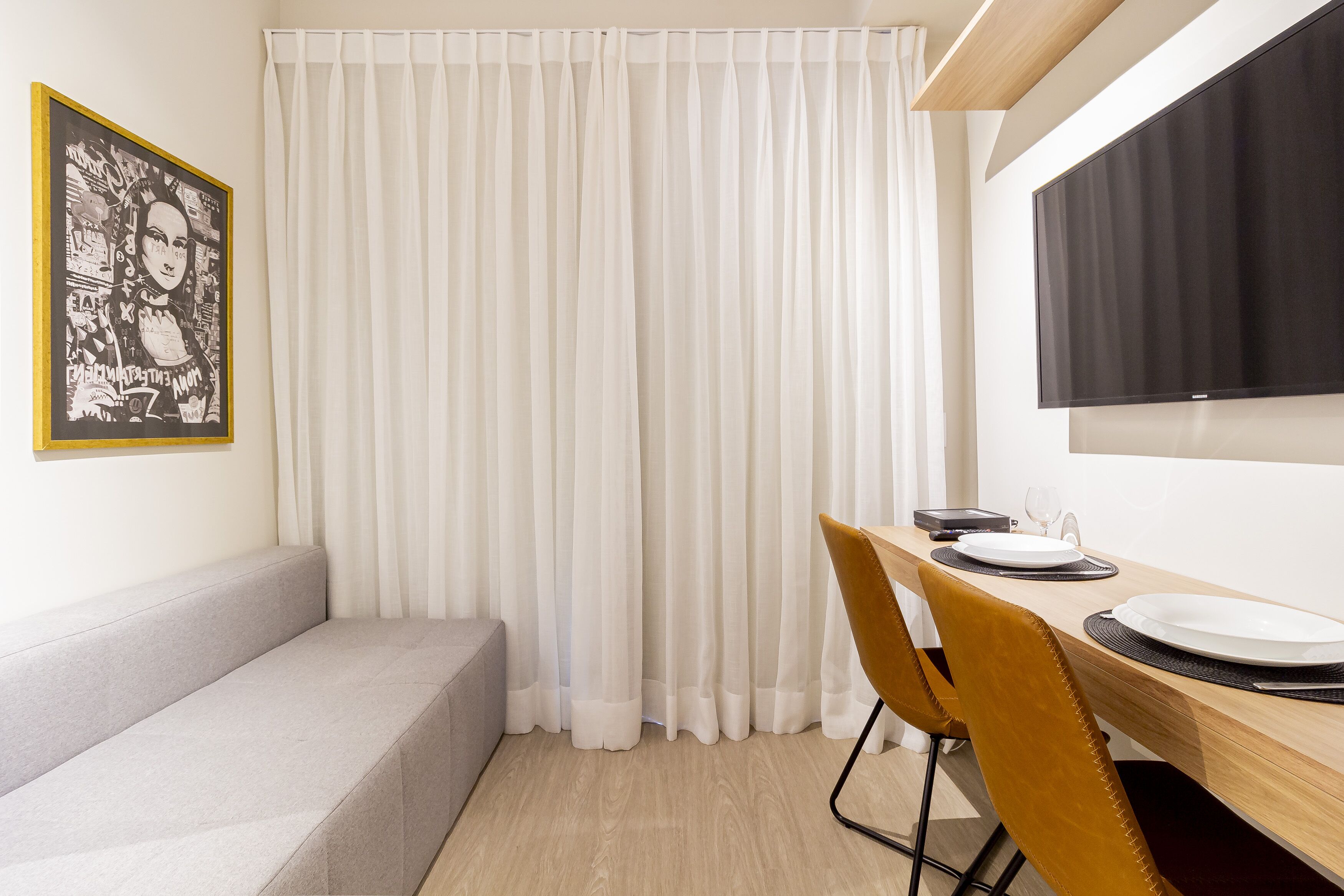 A modern room with a sofa, a dining table with chairs, a flat-screen TV, and a large window with white curtains.