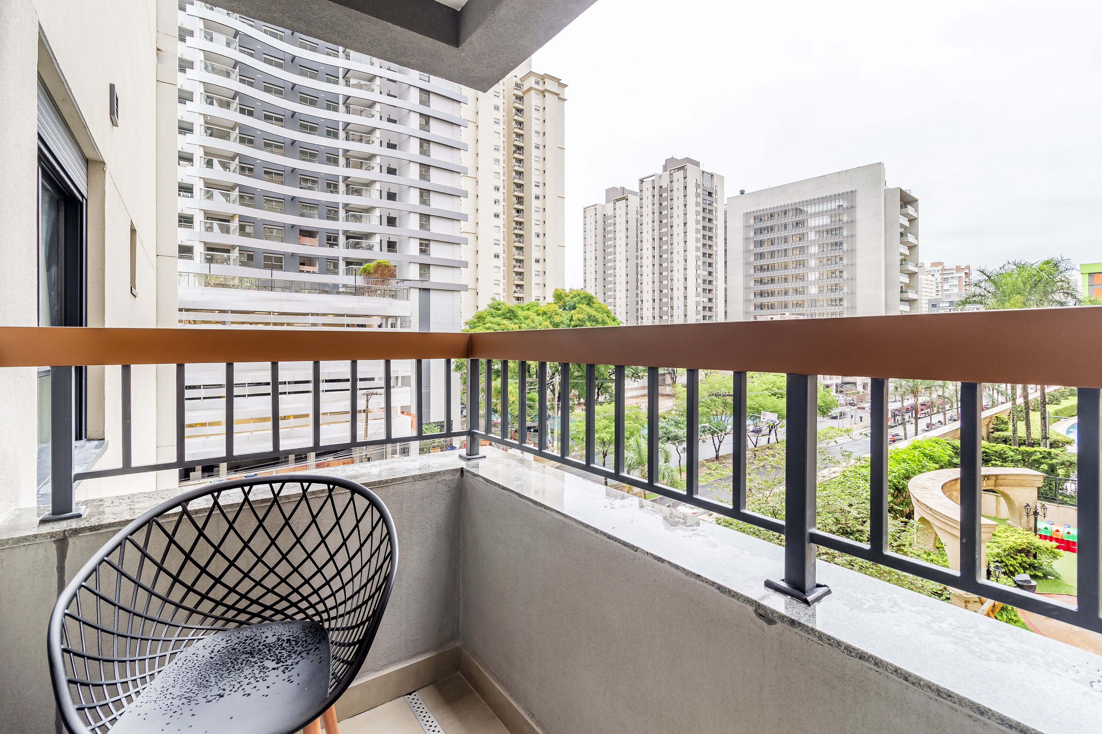 A balcony with a metal railing, a black wireframe chair, and a view of high-rise buildings and greenery.