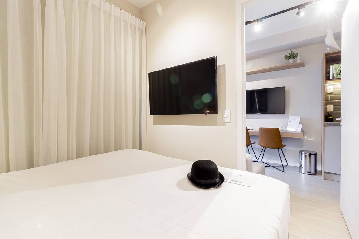 Ein modernes Hotelzimmer mit einem großen Bett, einem flachbildfernseher an der Wand, einem Schreibtisch mit Stuhl und einer kleinen Mülleimer.
