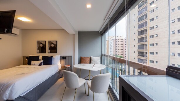 A modern hotel room with a bed, two chairs, a small table, and a large window offering a view of the city. - Charlie Uwin Brooklin (São Paulo)