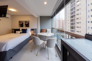 A modern hotel room with a bed, two chairs, a small table, and a large window offering a view of the city. - Charlie Uwin Brooklin (São Paulo)