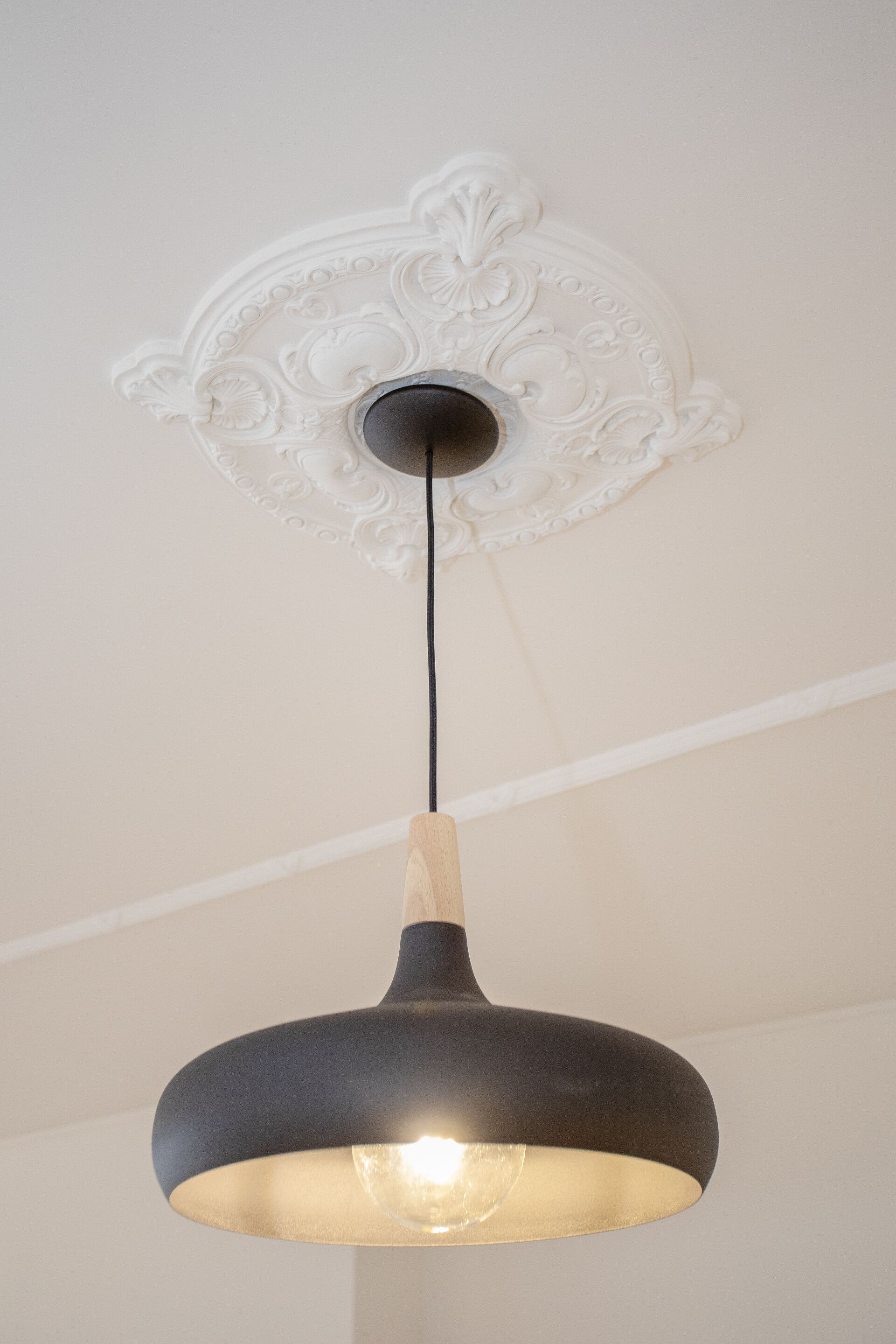 A pendant light fixture with a black shade and a wooden accent, hanging from a ceiling with an ornate white decorative panel.