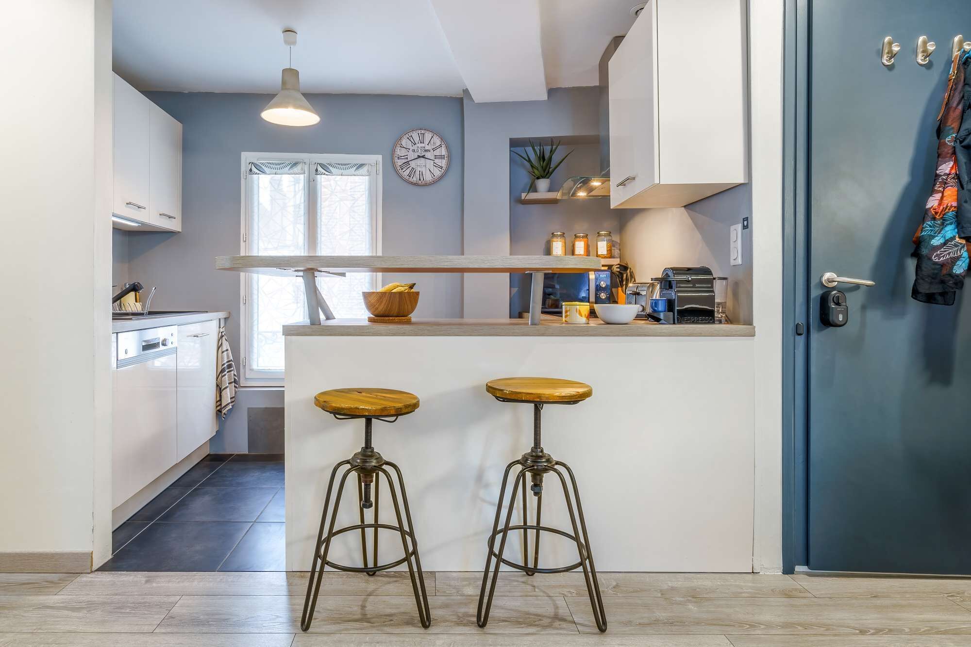Una cocina moderna con una isla central, dos taburetes, un reloj y una puerta con abrigos colgados.