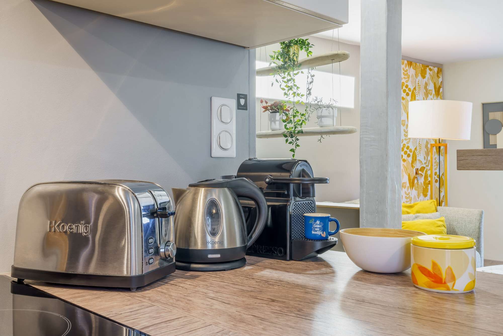 A kitchenette with a toaster, kettle, and coffee maker. There is a bowl, a mug, and a container on the counter.