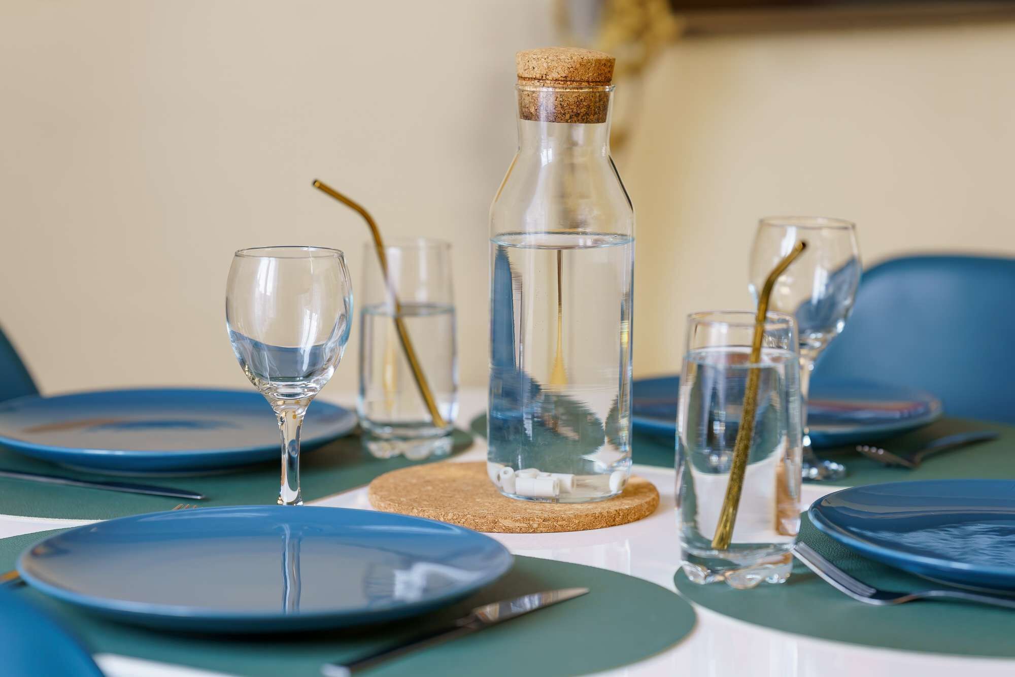 Une table à manger dressée avec des assiettes bleues, des verres transparents et une bouteille fermée par un bouchon.