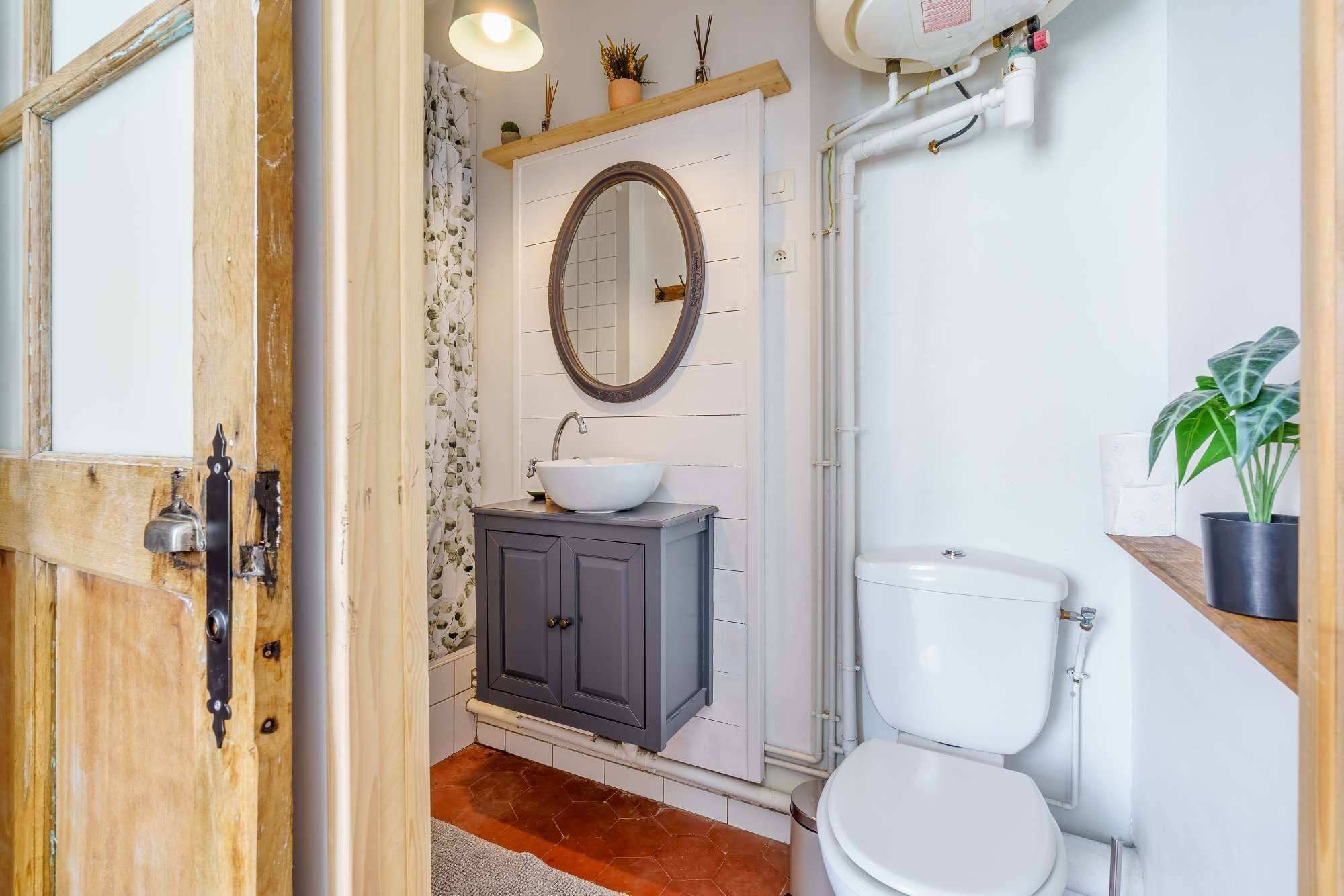 Une salle de bain avec une porte en bois, des toilettes blanches, un lavabo avec un receveur, un miroir et un espace douche.