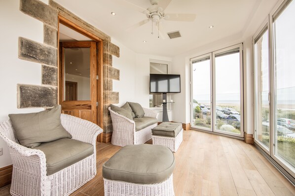 A spacious room with wicker furniture, a ceiling fan, and a large window offering a view of parked cars. - South View House (Alnwick)