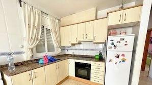 Una cocina con armarios de madera clara, horno empotrado, refrigerador con imanes y una ventana con cortinas.