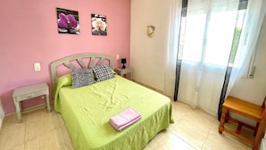 Un dormitorio con cama, mesita de noche, banco de madera, ventana con cortinas y dos cuadros colgados en la pared.