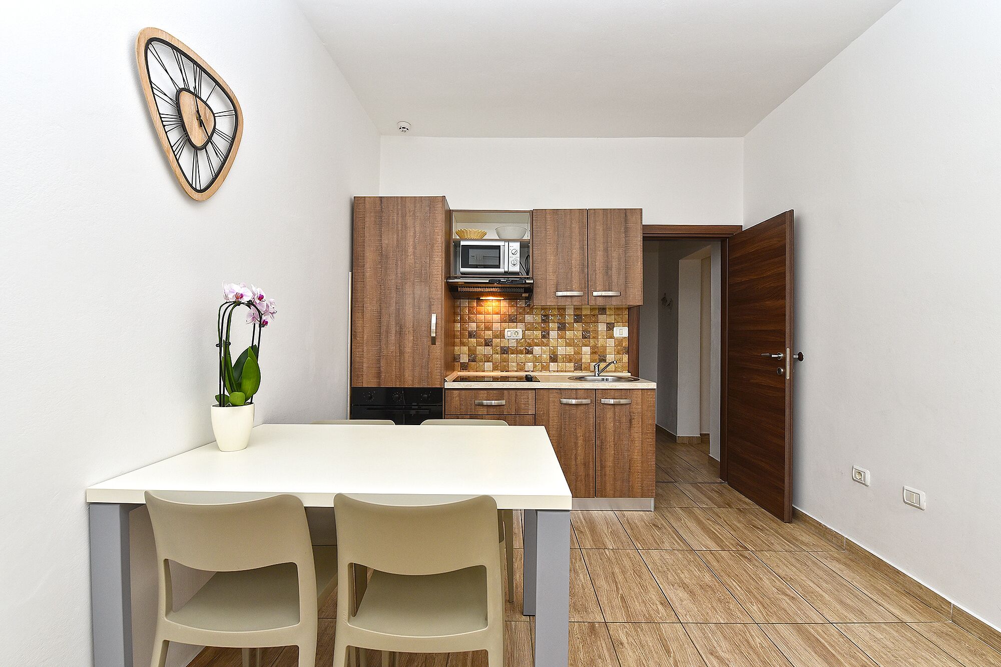 Een kleine keuken met houten kastjes, een magnetron en een betegelde spatwand. Er staat een eettafel met stoelen en een potorkidee.