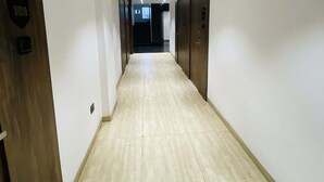 A hallway with a light wooden floor, white walls, and a door with a number plaque. - Hotel Vijay Shree (Sohagpur)