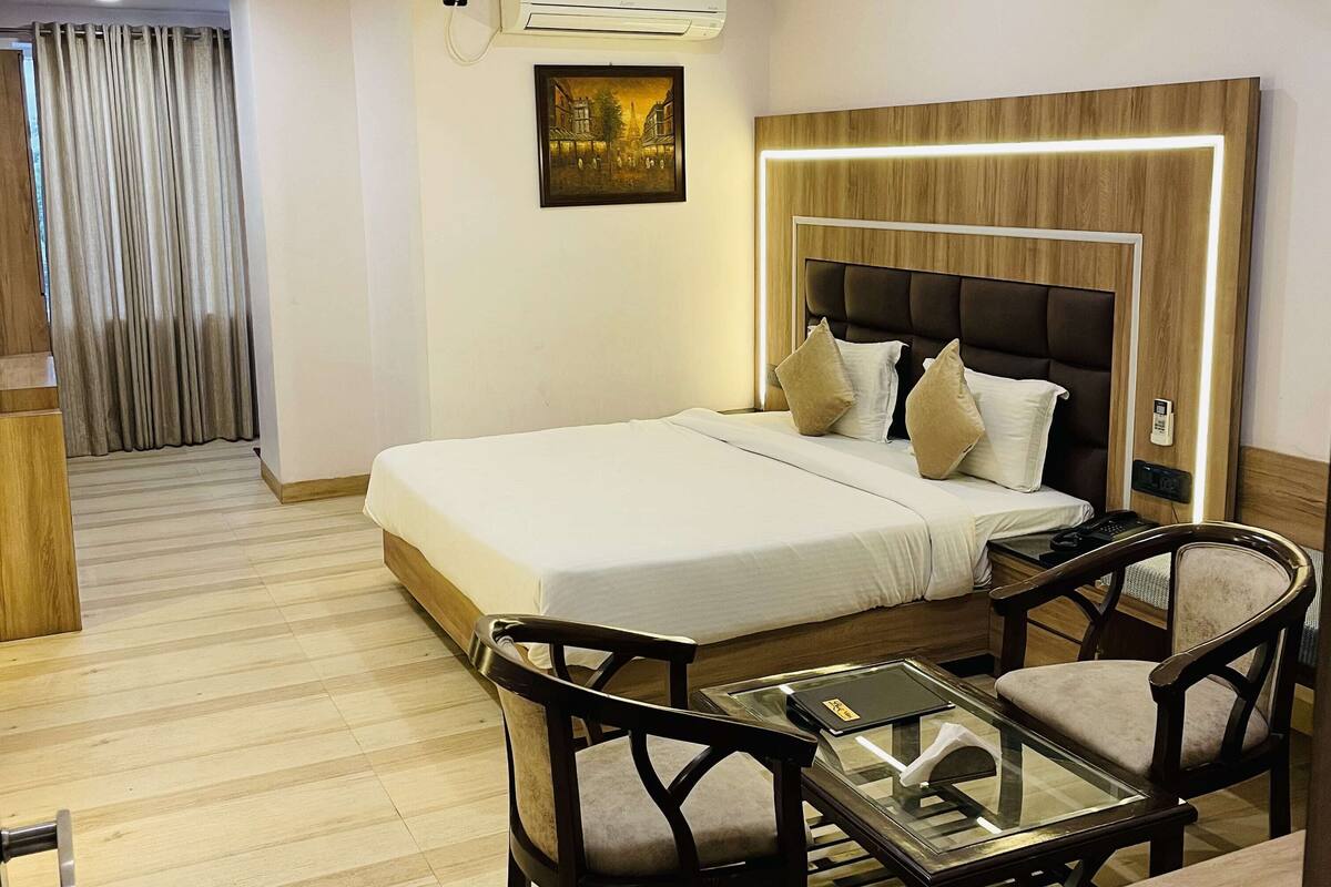 A hotel room with a bed, bedside table, chairs, a glass table, an air conditioning unit, and a painting on the wall.