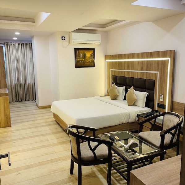 A hotel room with a bed, bedside table, chairs, a glass table, an air conditioning unit, and a painting on the wall.