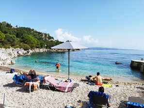 A pebble beach with clear blue water, a rocky shoreline, and a few people sunbathing.