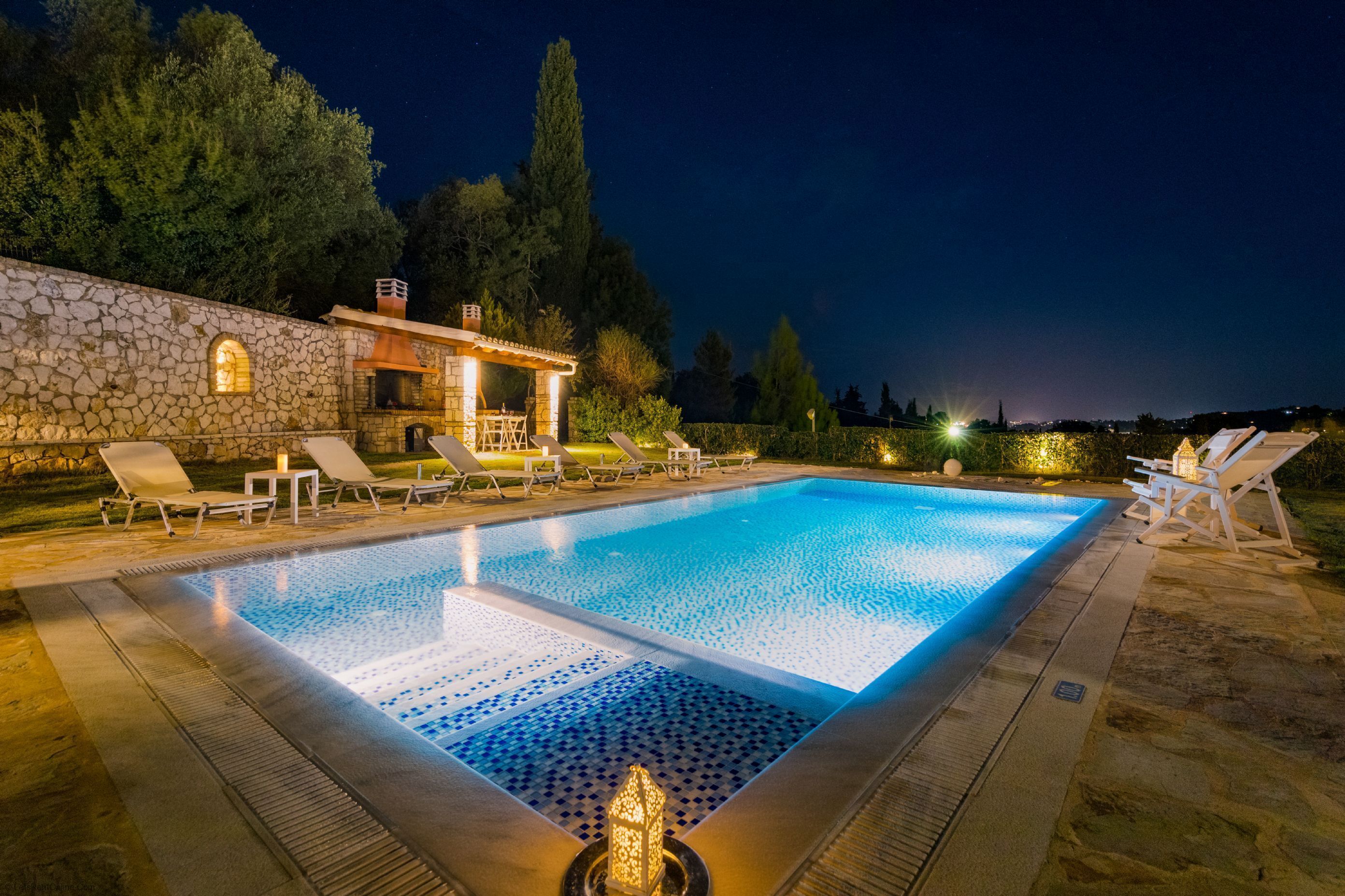 A rectangular swimming pool with a stone wall and lounge chairs on one side.