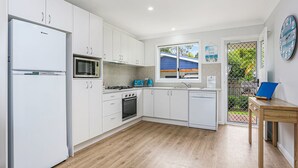 Een moderne keuken met witte kasten, een koelkast, een magnetron, een fornuis en een vaatwasser. Er is een raam met uitzicht op een blauw huis en een tuin.