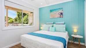 Een slaapkamer met een groot bed, een raam met uitzicht op groen, een schilderij van een strand en een klein nachtkastje met een lamp.