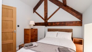 A bedroom with a wooden ceiling, a bed with white bedding, a nightstand, a wooden dresser, and a door. - The Stables (Colne)