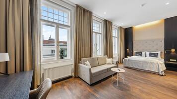 Ein modernes Hotelzimmer mit einer Couch, einem Bett, einem kleinen Tisch und Blick auf die Stadt durch große Fenster.