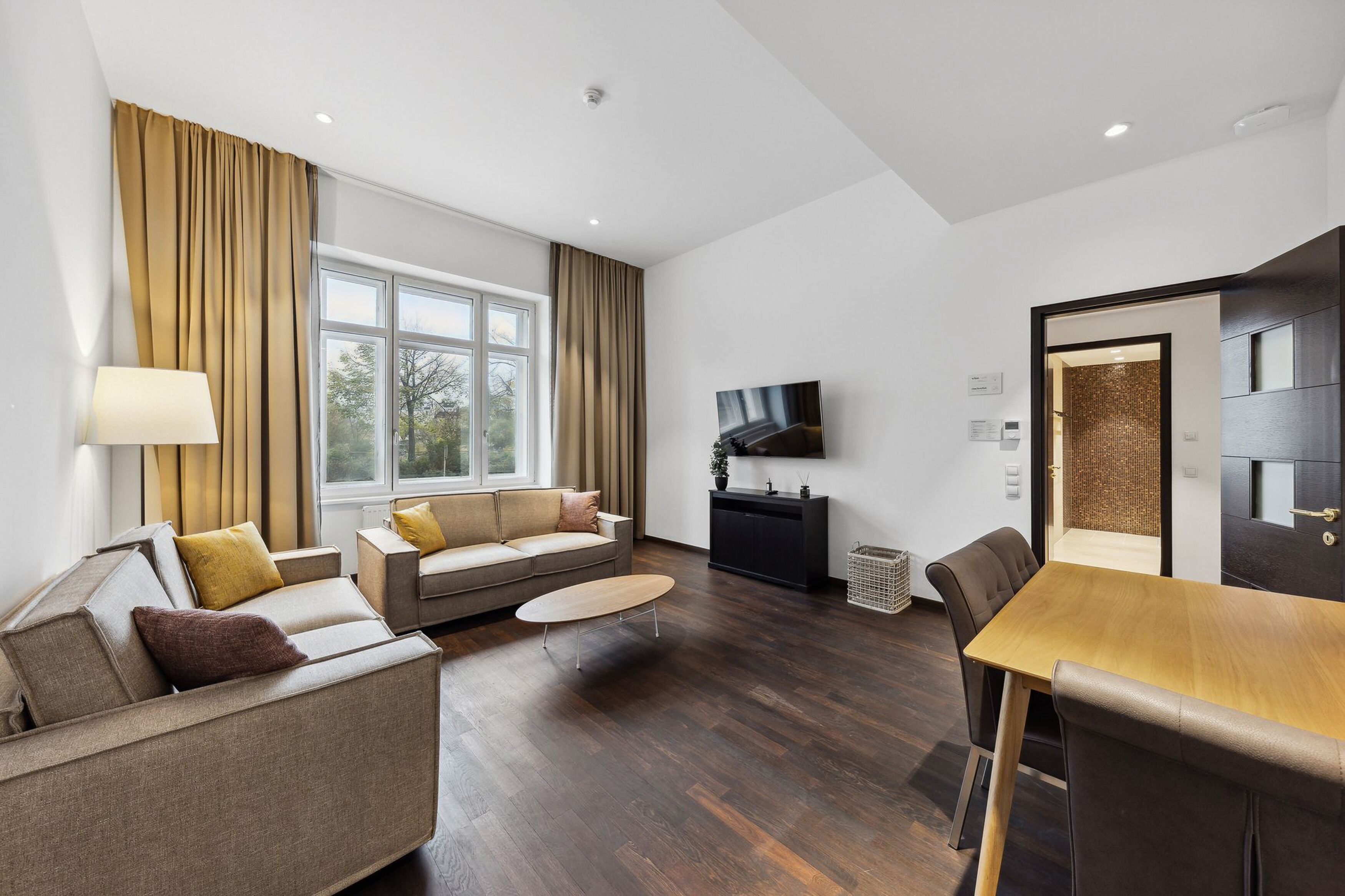 Een moderne woonkamer met een zithoek, een salontafel en een eettafel. Er is een televisie aan de muur bevestigd en een raam met gordijnen.