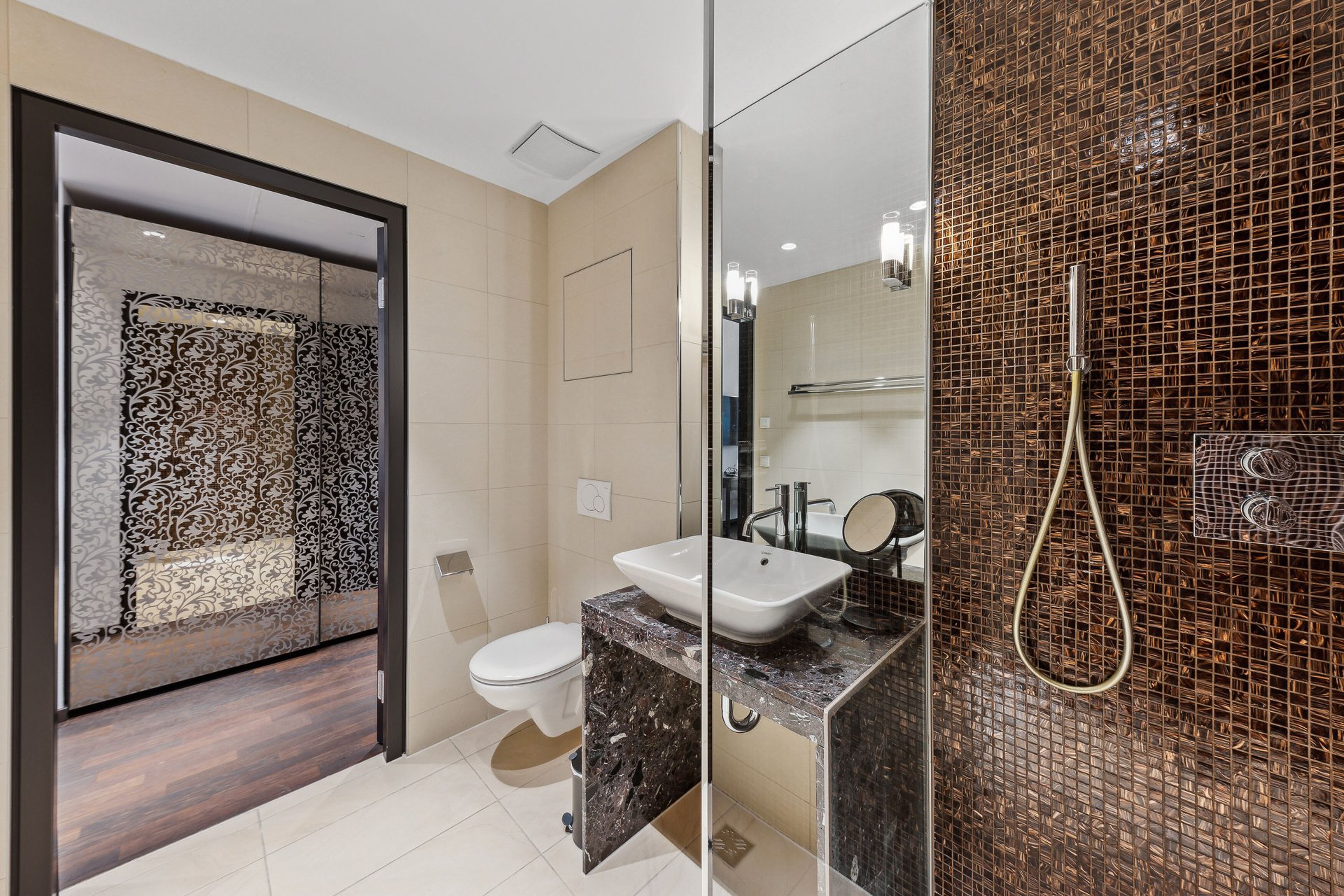 A modern bathroom with a glass partition, a white sink, a toilet, and a shower area with a mosaic wall.