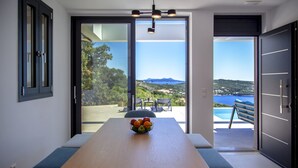 A modern dining area with a wooden table, a bowl of fruit, and a view of a pool and landscape. - Villa Blauw with Infinity Pool  (LEFKADA)