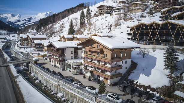 A snow-covered mountain town with a hotel featuring multiple floors, balconies, and a ski lift visible in the background. - Hotel Forellenhof (Saalbach-Hinterglemm)