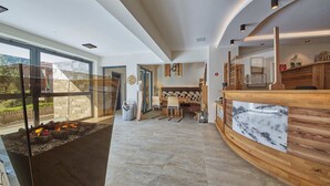 A modern interior with a stone fireplace, a wooden reception desk, and a seating area with a view of the outdoors. - Hotel Forellenhof (Saalbach-Hinterglemm)