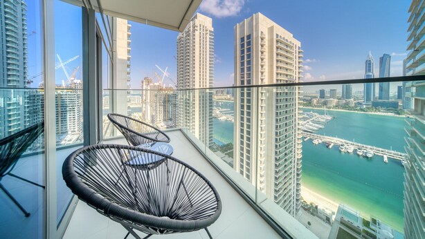 A balcony with two black wicker chairs, a glass railing, and a view of a marina with boats and high-rise buildings.