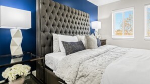 A bedroom with a tufted headboard, a large bed with white and gray bedding, two bedside tables with lamps, and a window with blinds. - Exquisite Apt near Beaumont (Royal Oak)